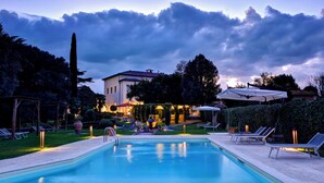 En swimmingpool med liggestole og et pergola-område, omgivet af en have og en bygning med tegltag.