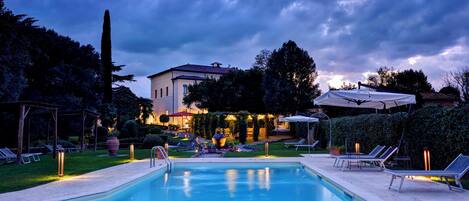 A swimming pool with lounge chairs and a pergola, surrounded by a garden and a building with a tiled roof.