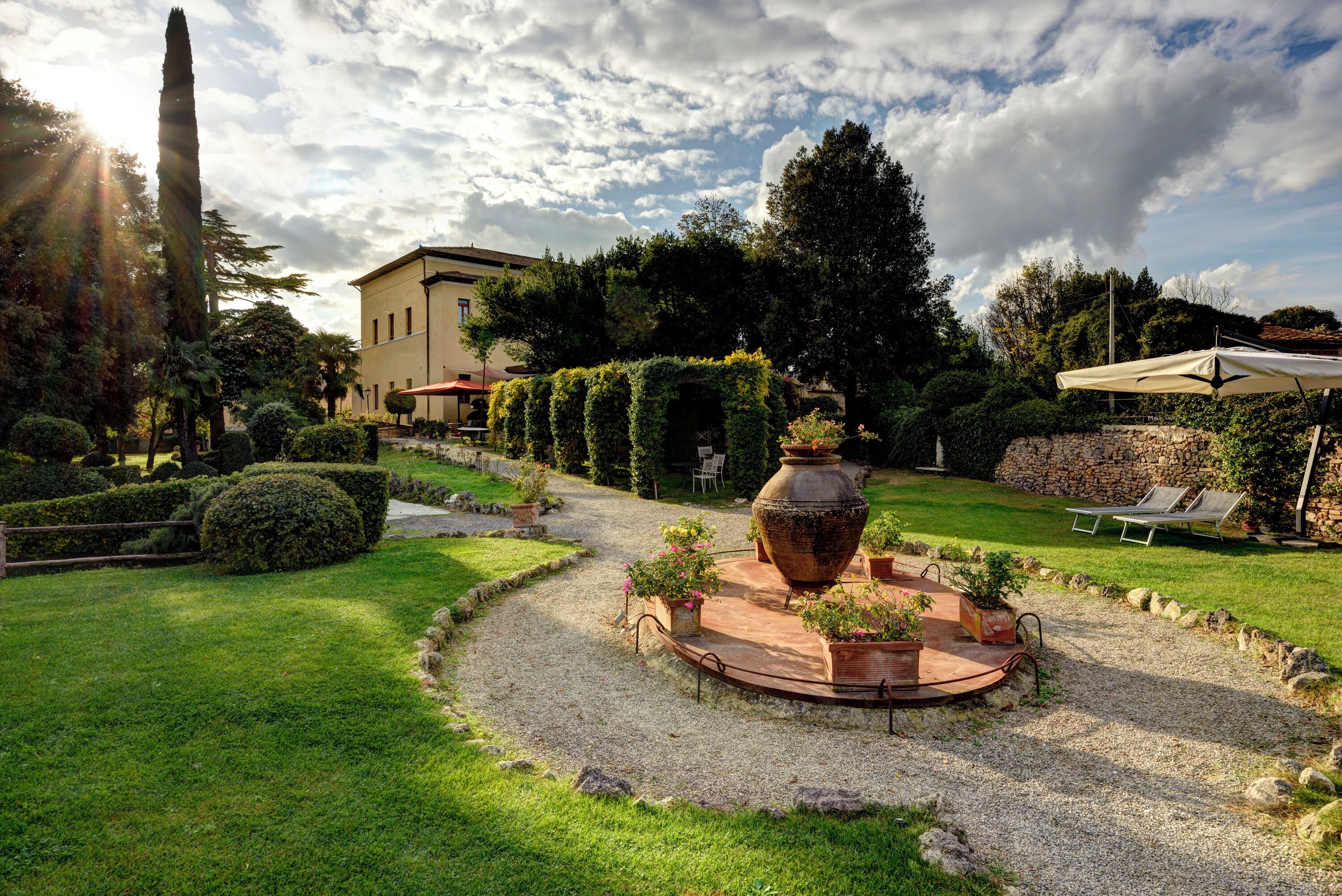 Un jardín con un patio circular, una urna grande y un edificio al fondo.