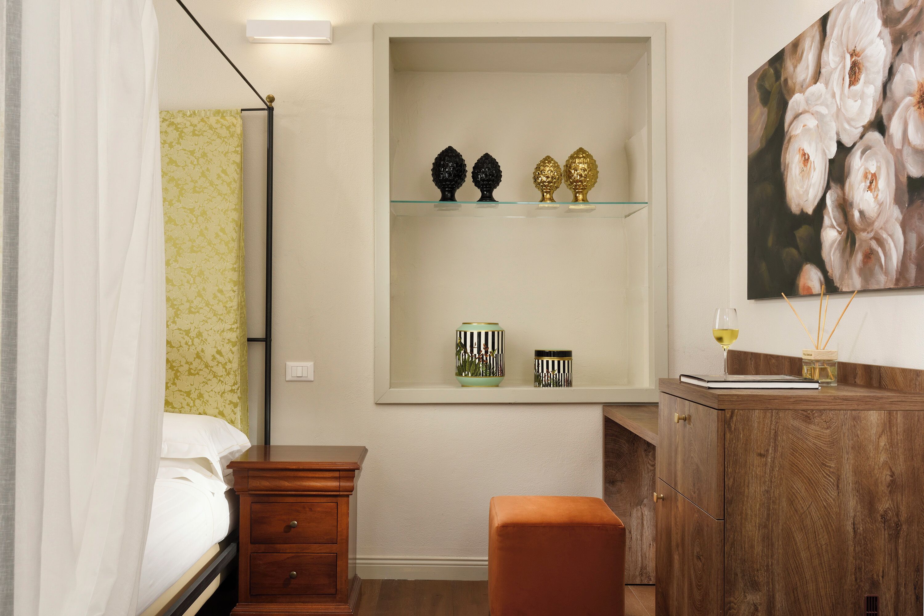 Een slaapkamer met een bed, een houten nachtkastje, een dressoir, een spiegel en een plank met decoratieve objecten.