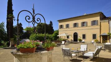 Um pátio externo com mesas e cadeiras de pedra, rodeado por plantas em vasos e uma peça decorativa em metal.