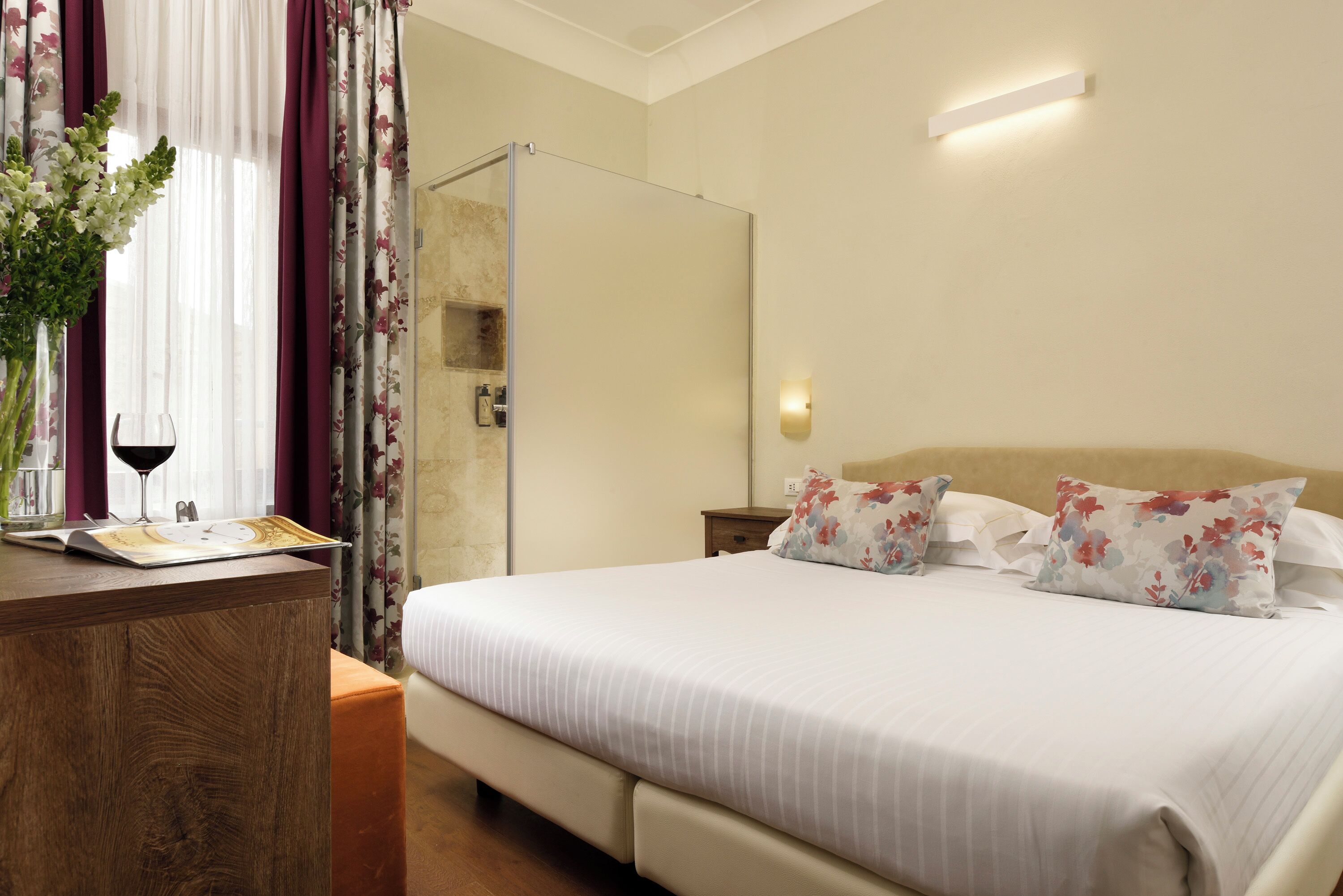 Una camera d'albergo con un letto, una scrivania in legno, un vaso con fiori e una finestra con tende.