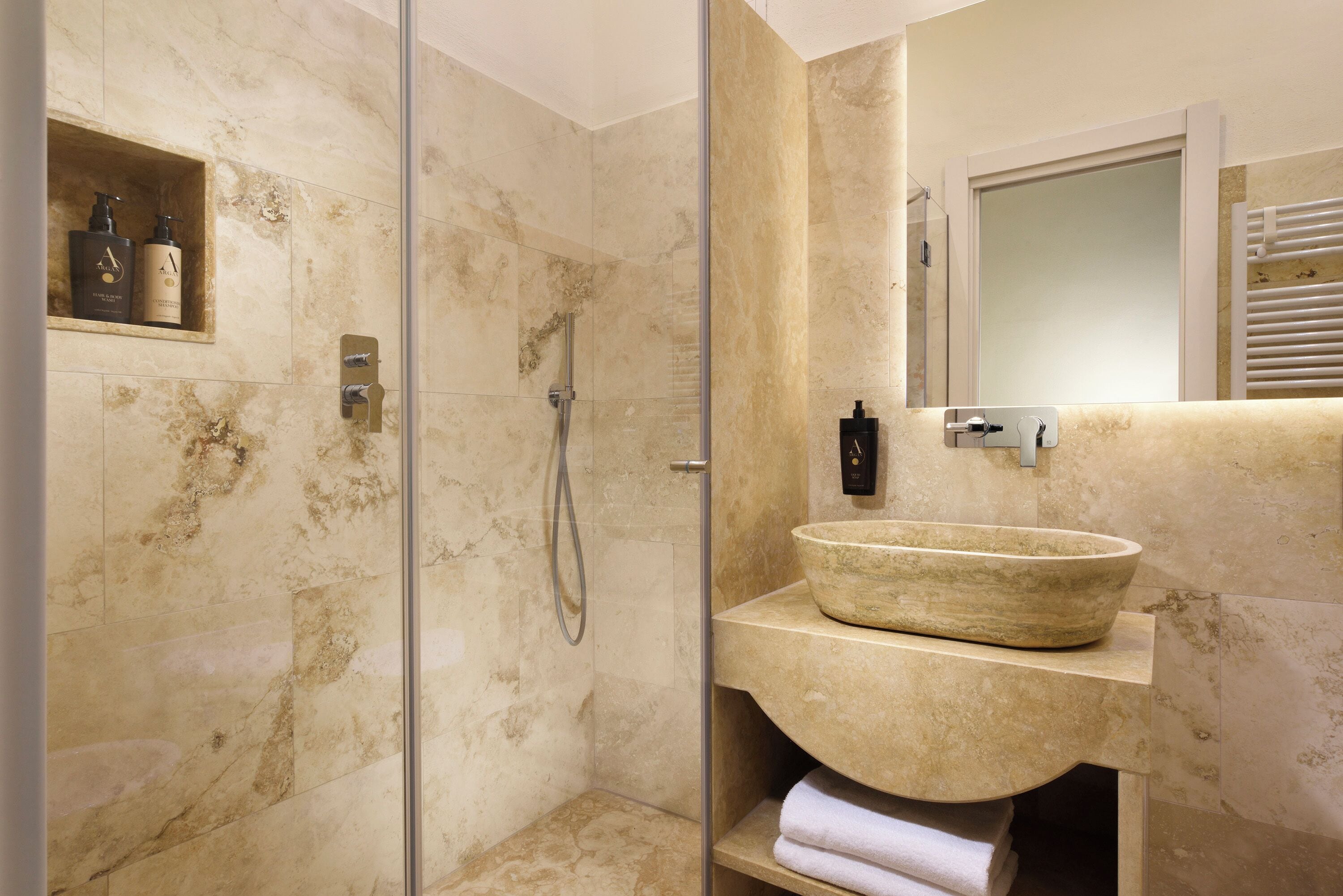 A bathroom with a glass-enclosed shower, a marble sink, and a shelf with toiletries.