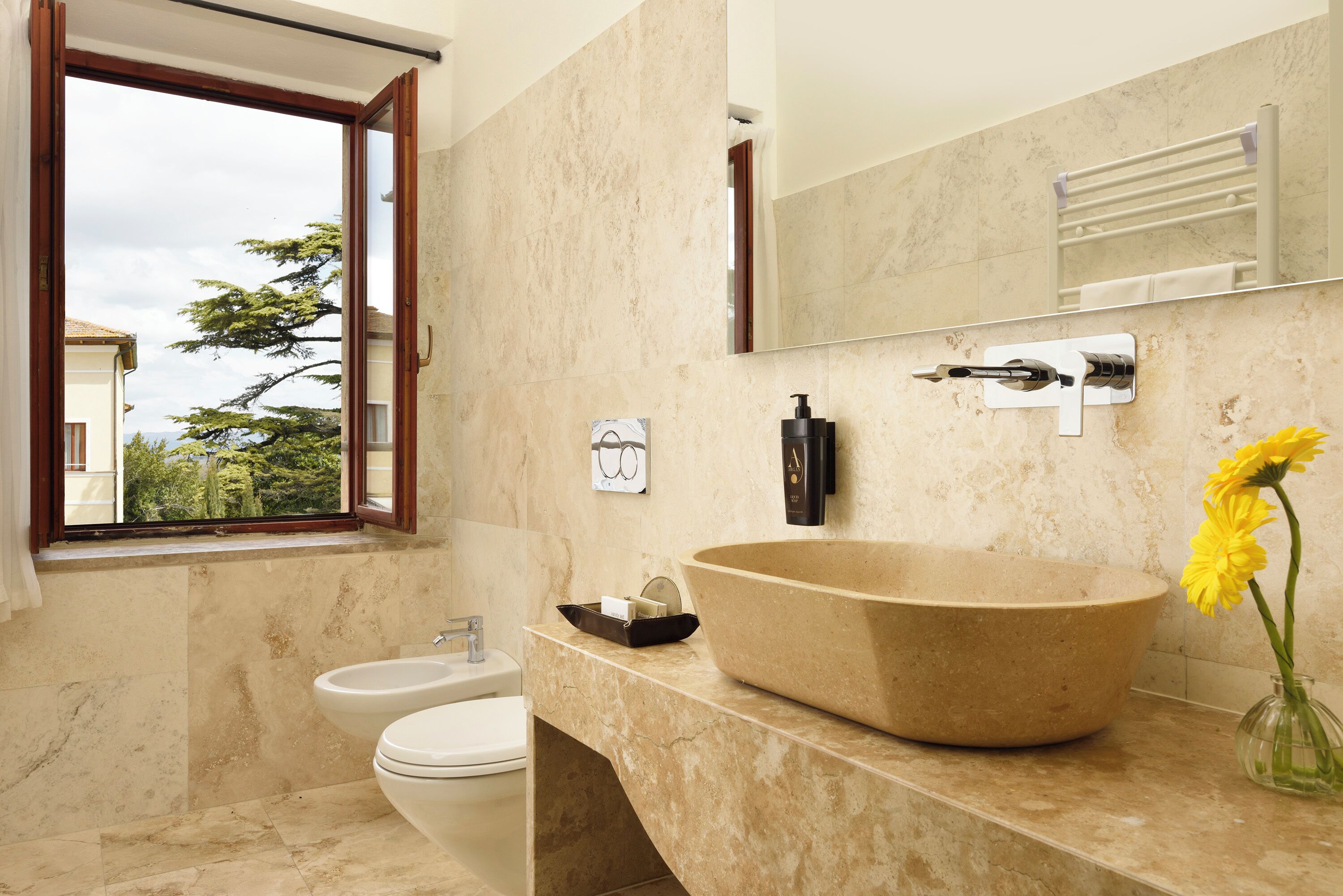 A bathroom with a large beige sink, a bidet, a window with a view of trees, and a mirror above the sink.