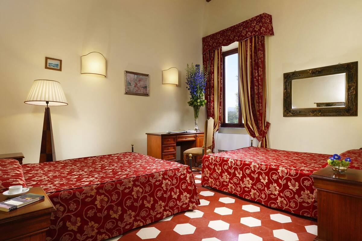 A hotel room with two beds, a desk, a chair, a lamp, a vase with flowers, a mirror, and a window with curtains.