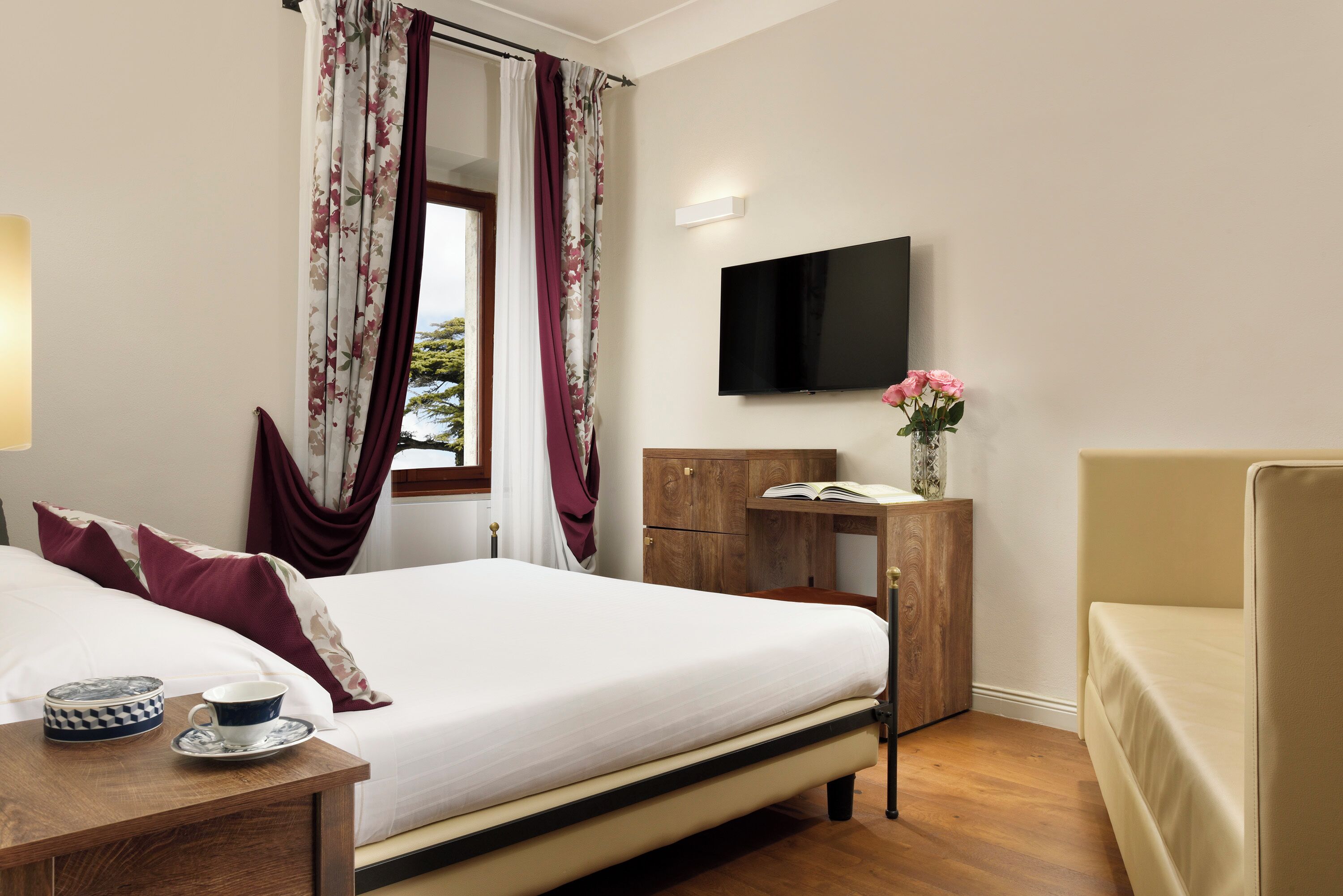 Une chambre d’hôtel avec un lit, une table de chevet en bois sur laquelle se trouvent une tasse et une soucoupe, un bureau avec un livre et un vase de fleurs, un téléviseur fixé au mur et une fenêtre avec des rideaux.