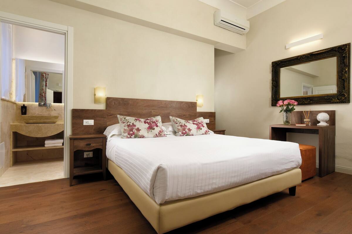 A bedroom with a wooden headboard, a bedside table with a lamp, a mirror, and a vanity with a sink.