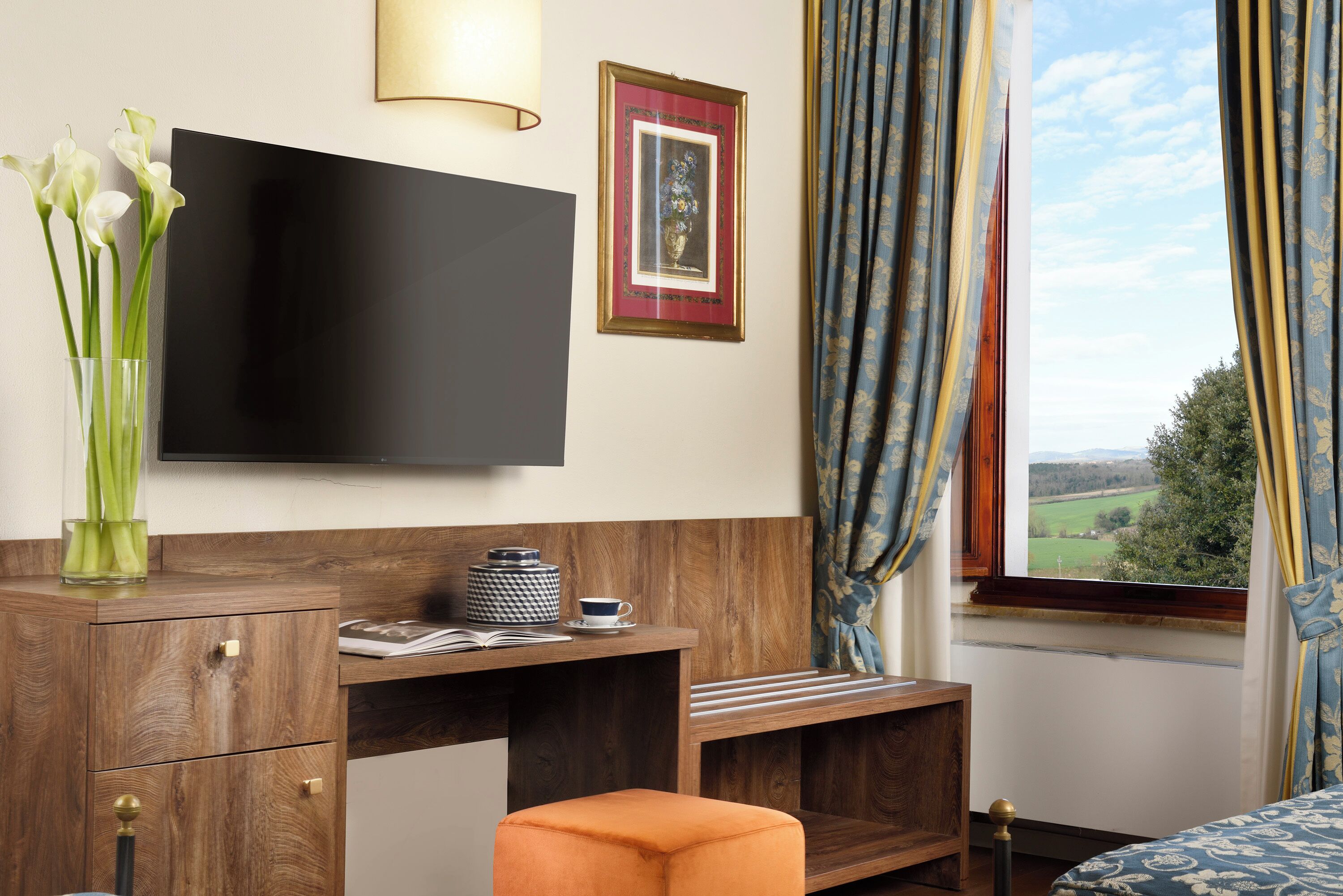Une chambre d’hôtel équipée d’un téléviseur à écran plat, d’un bureau en bois, d’un banc rembourré, d’un vase avec des fleurs et d’une fenêtre avec des rideaux.