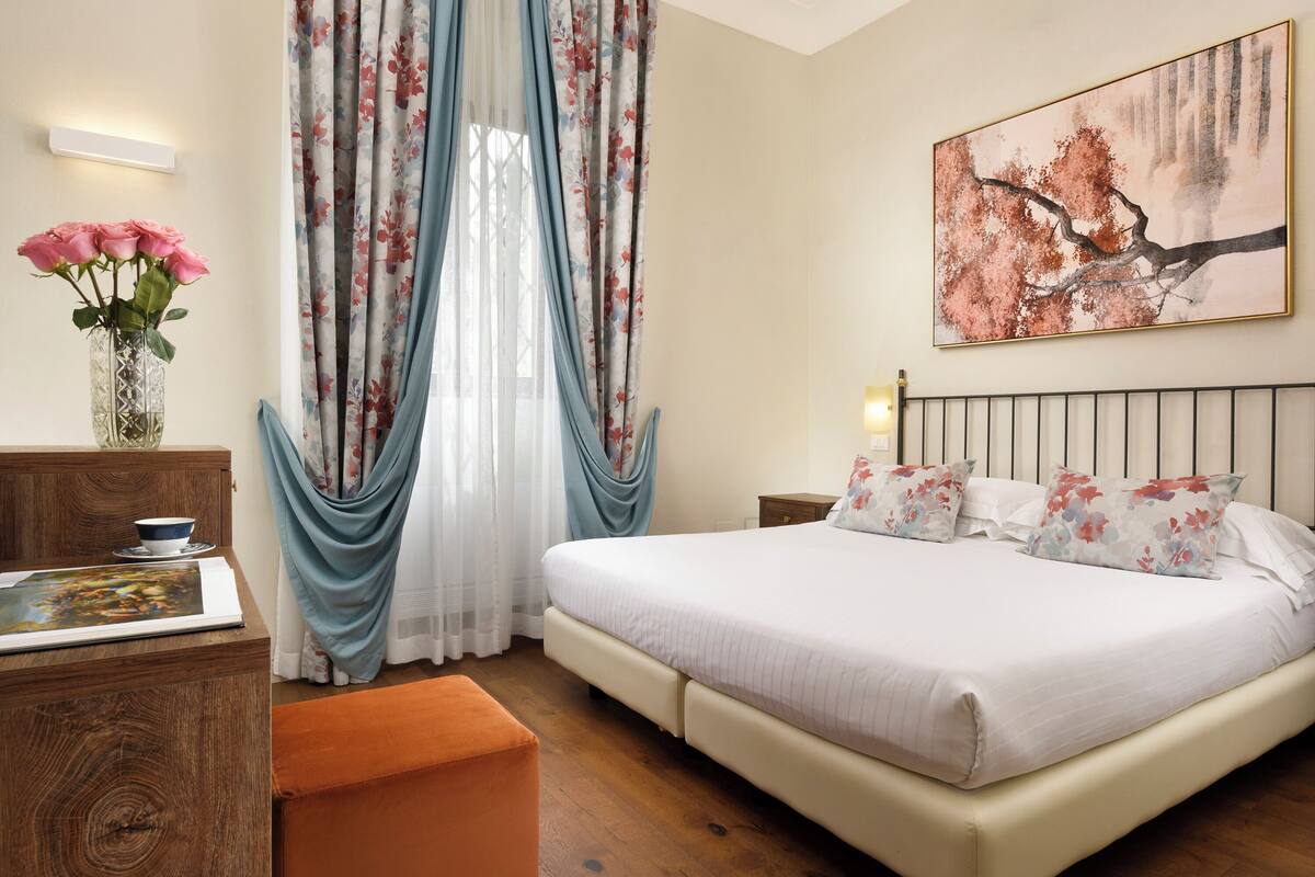 A bedroom with a bed, a nightstand, a bedside table, a vase with flowers, and a painting on the wall.