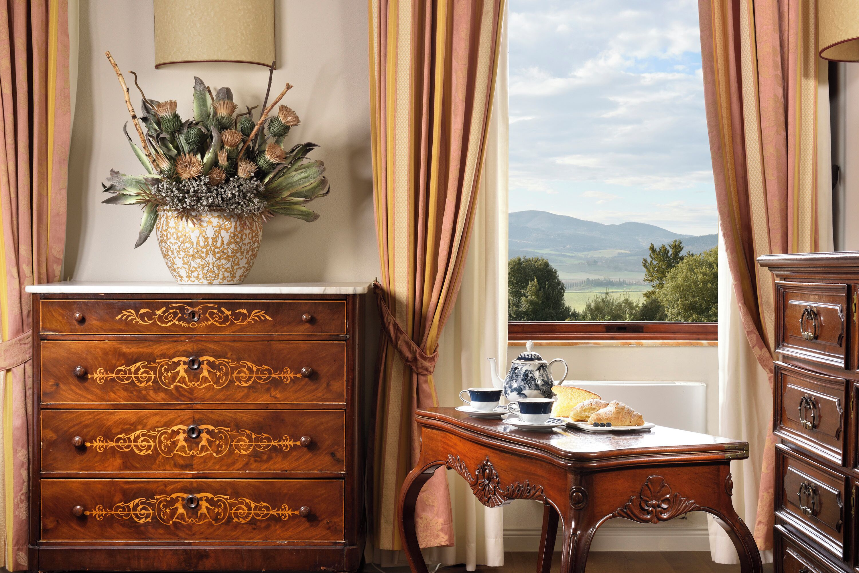 A room with a wooden dresser, a table with a teapot and cups, and a view of the countryside through a window.