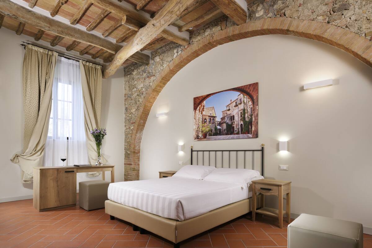 A bedroom with a bed, bedside tables, a desk, a window with curtains, and a painting on the wall.