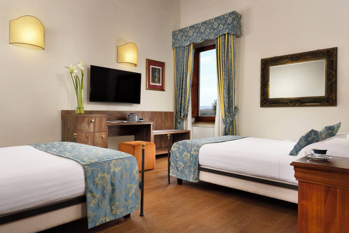 A hotel room with two beds, a desk, a television, and a large window with curtains.