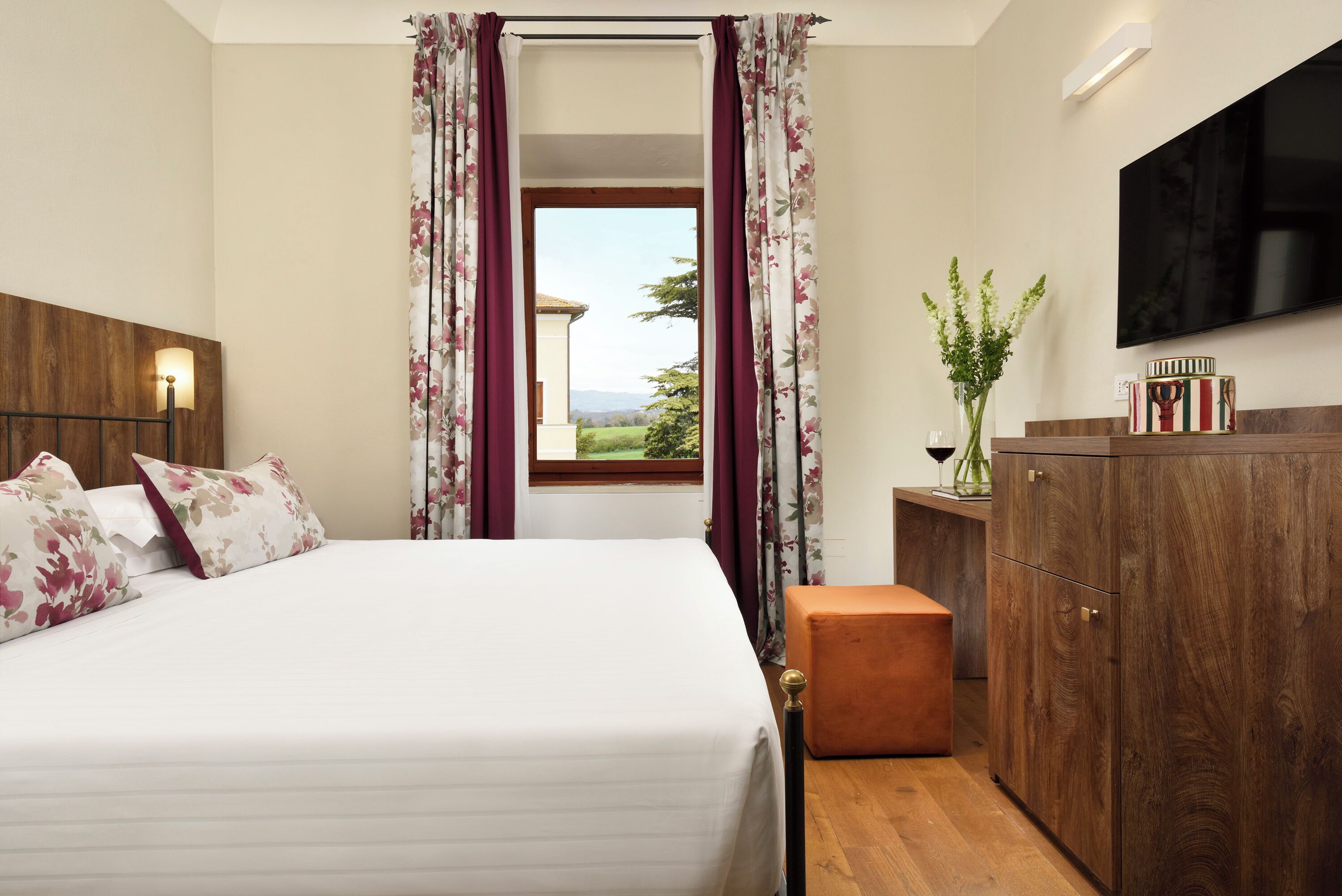 A bedroom with a bed, a wooden nightstand, a TV mounted on the wall, and a window with curtains.