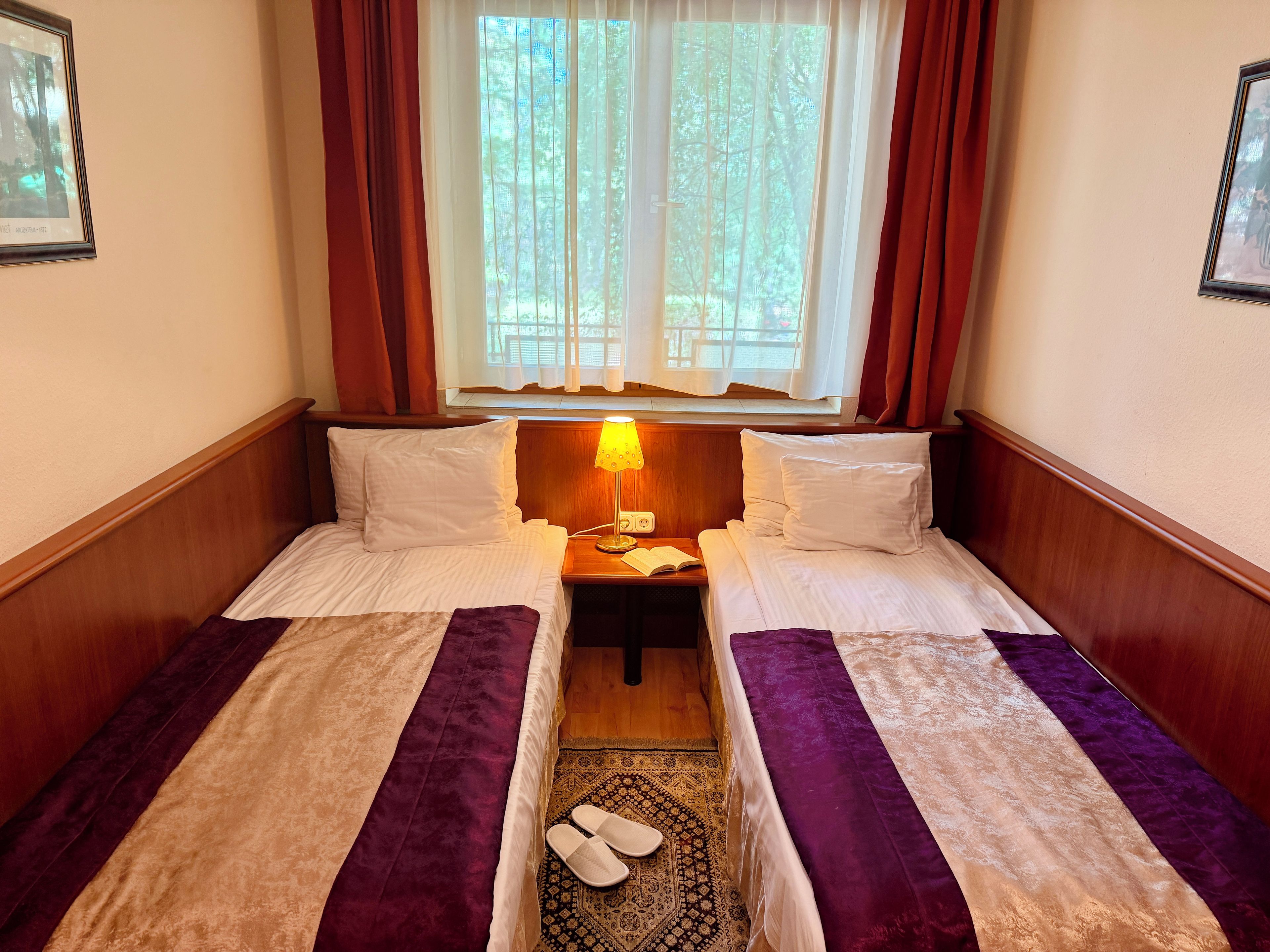 A hotel room with two beds, a nightstand, a lamp, and a window with curtains.