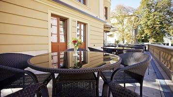 A patio with wicker furniture and a glass table, overlooking a street with trees and a building.