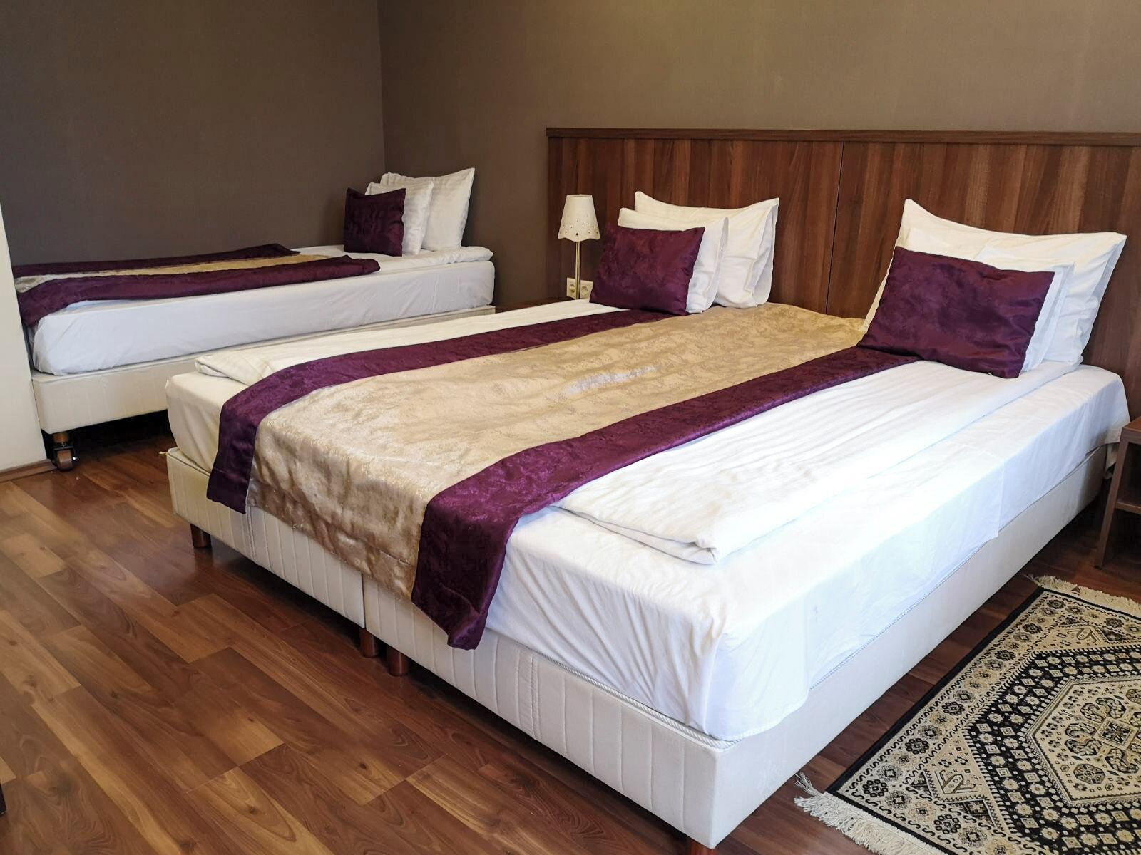 A hotel room with two beds, a wooden headboard, a patterned rug, and a nightstand with a lamp.
