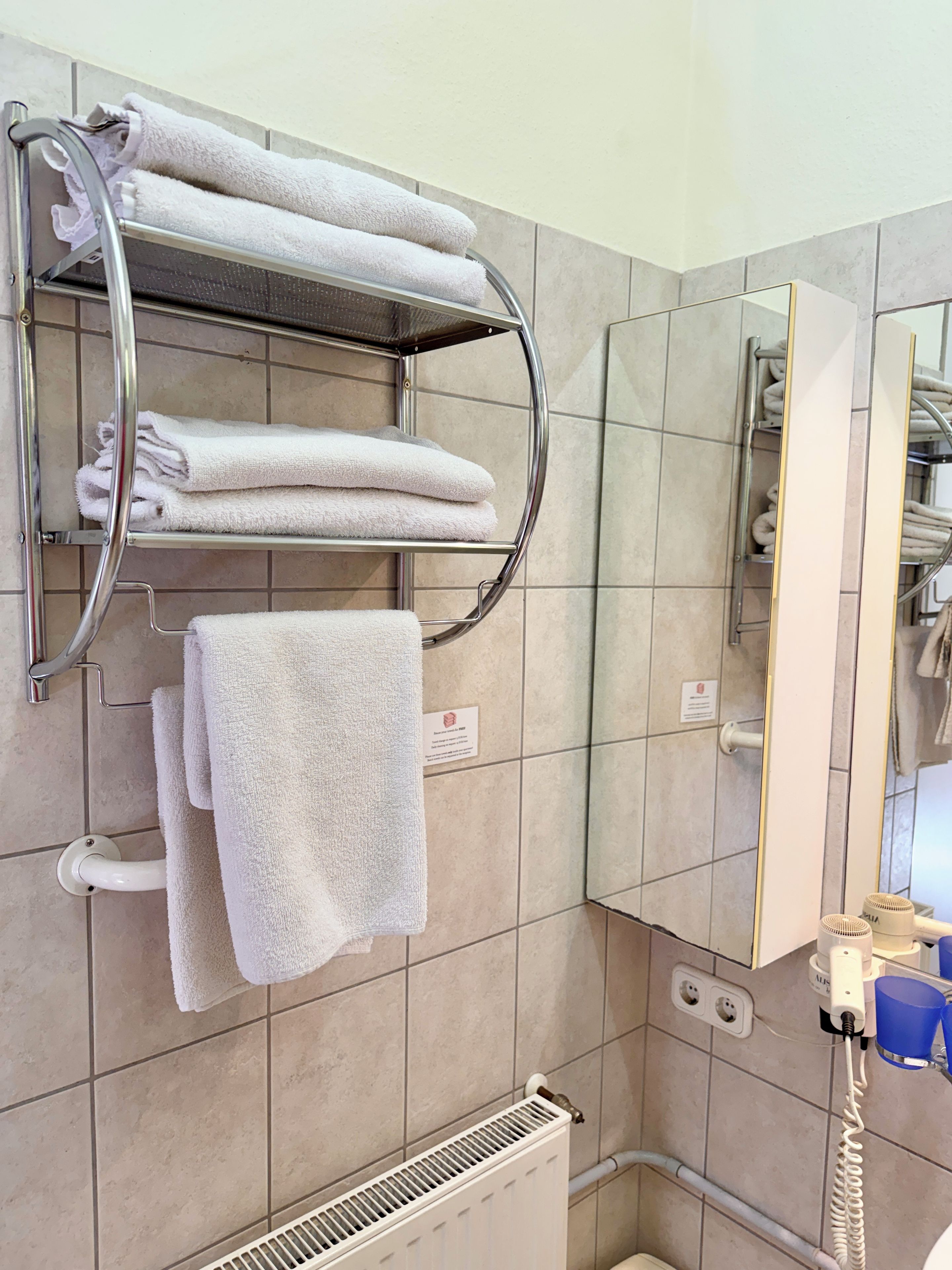 Un bagno con un portasciugamani pieno di asciugamani piegati, uno specchio, un termosifone e un asciugacapelli sul ripiano.