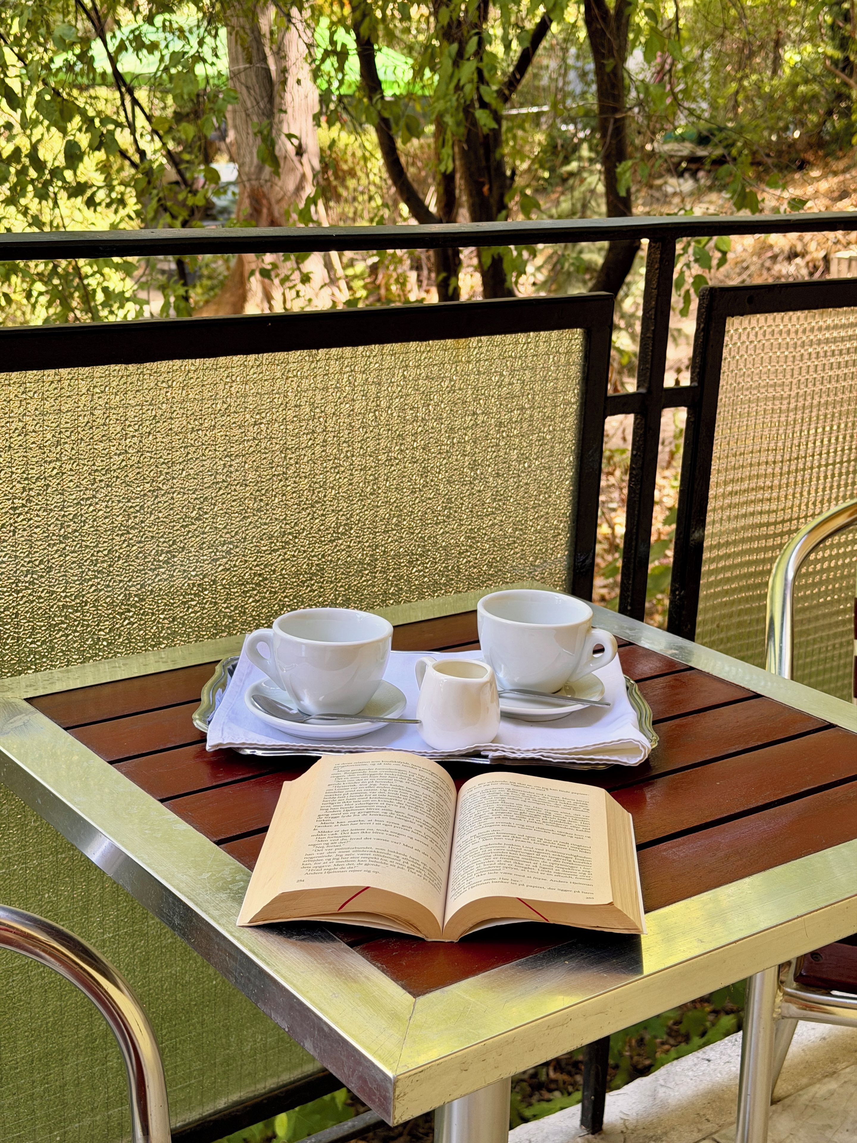 An outdoor table with a book, two cups, and a small pitcher, set against a backdrop of trees.