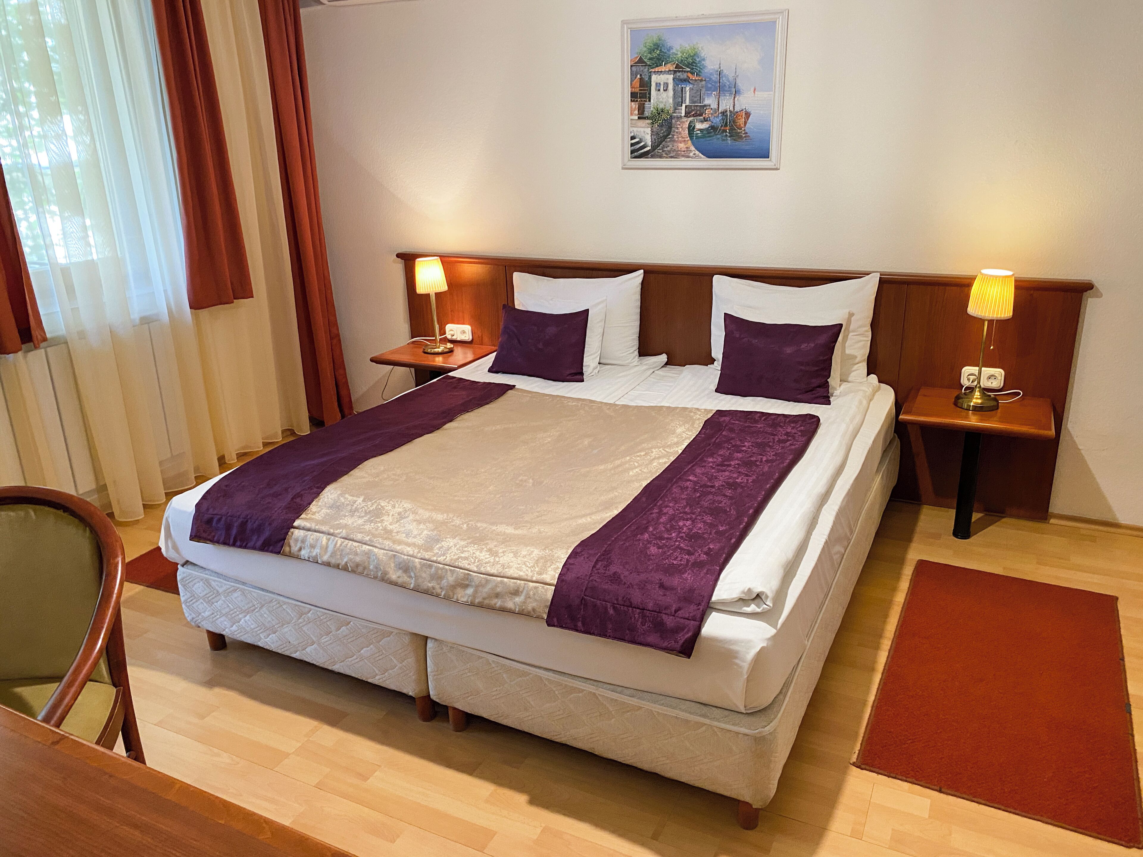 A hotel room with a large bed, two bedside tables with lamps, a chair, a painting on the wall, and a wooden floor.