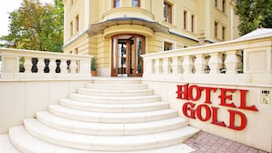 L’entrée d’un hôtel, avec un grand escalier menant à une porte, et une enseigne indiquant « HÔTEL GOLD ».