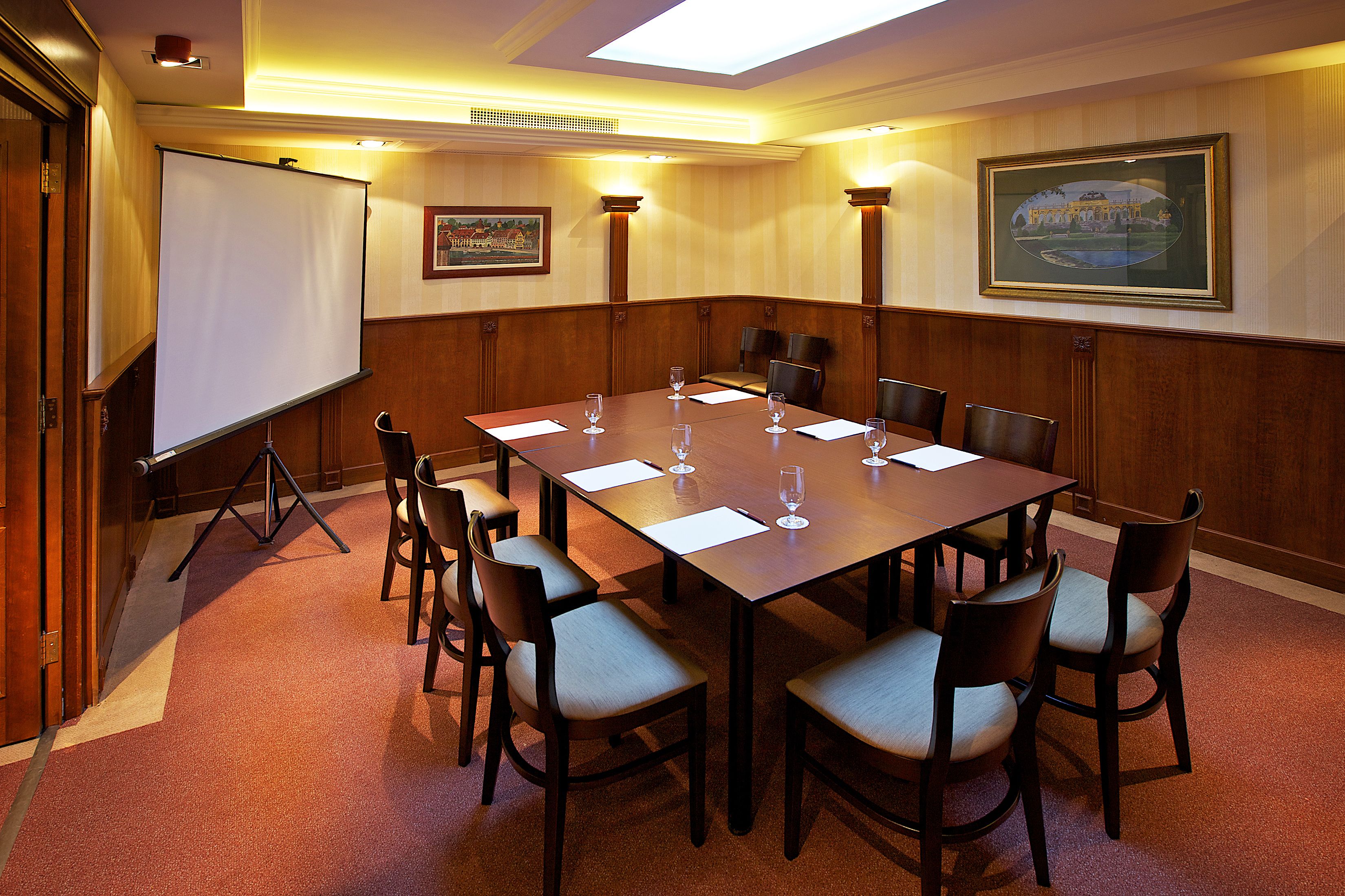 Una sala de conferències amb una taula gran, cadires, una pantalla de projector i quadres amb marc a les parets.