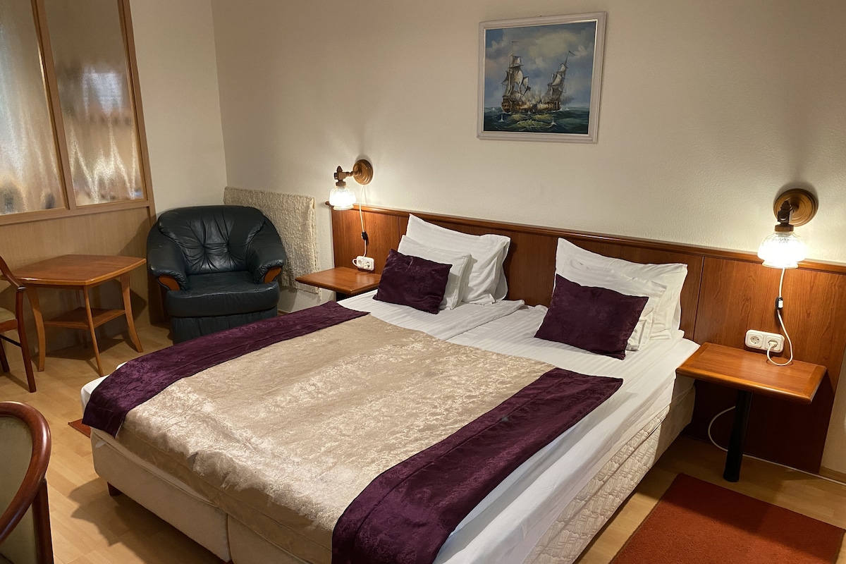 Una habitación de hotel con una cama grande, dos mesitas de noche con lámparas, una silla, una pequeña mesa y un cuadro en la pared.