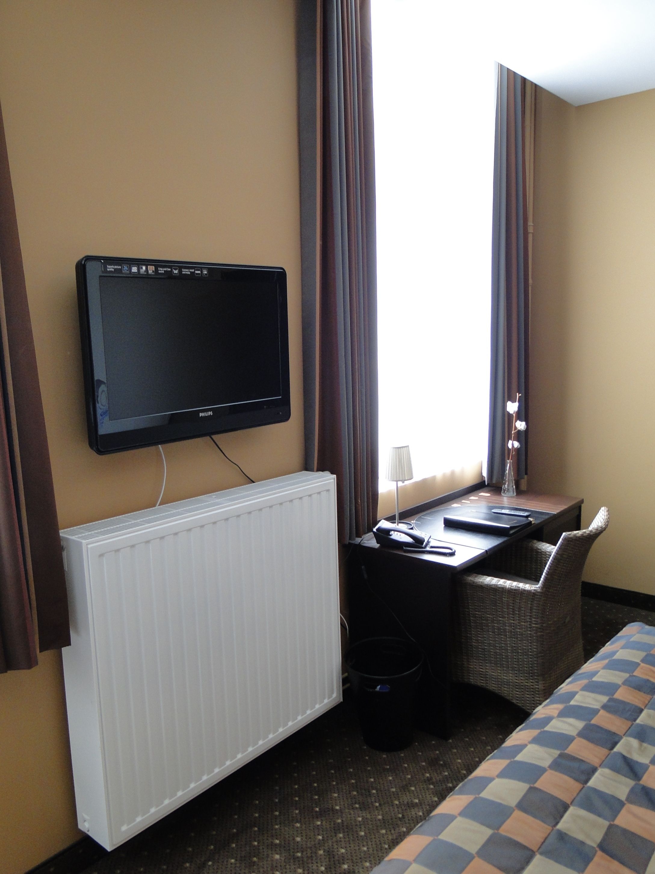 A hotel room with a flat-screen TV mounted on the wall, a desk with a chair, a radiator, and a bed with a checkered bedspread.