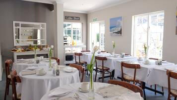 Un área de comedor con mesas redondas, manteles blancos y sillas de madera. Hay señalización para una cafetería y una salida.