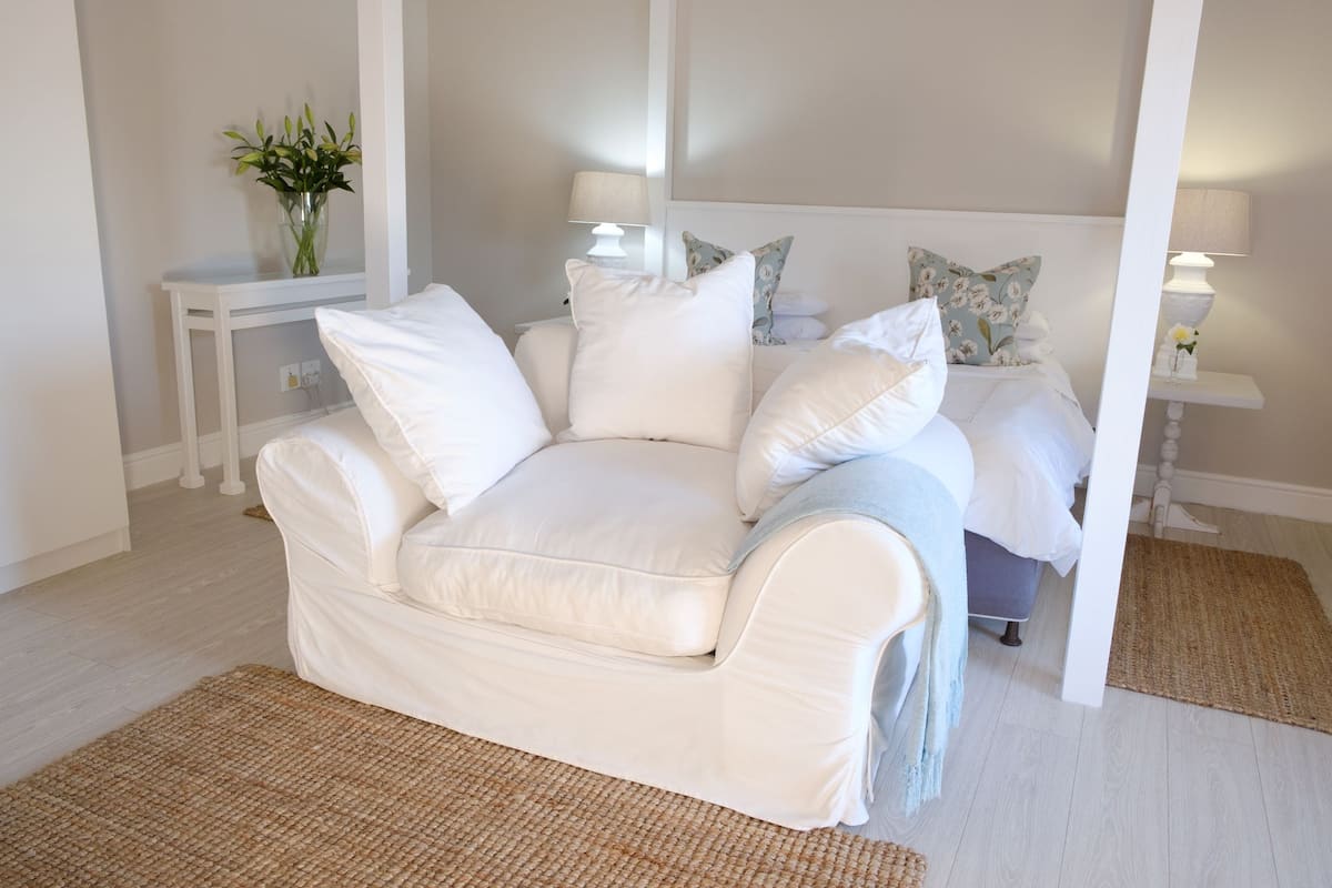 Un dormitorio con un sofá blanco, una cama con almohadas florales, una mesita de noche con una lámpara y un florero con flores.