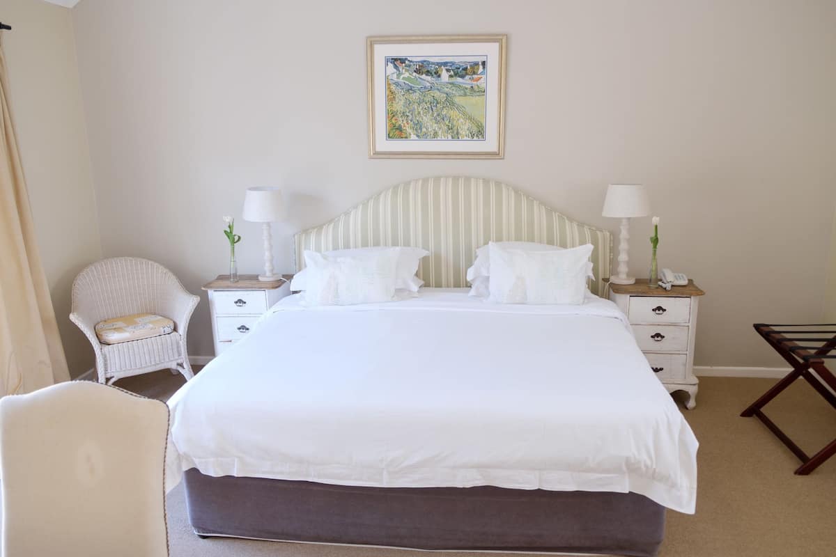 Un dormitorio ordenado con una cama grande, dos mesitas de noche con lámparas, una silla de mimbre y una pequeña mesa con un jarrón.