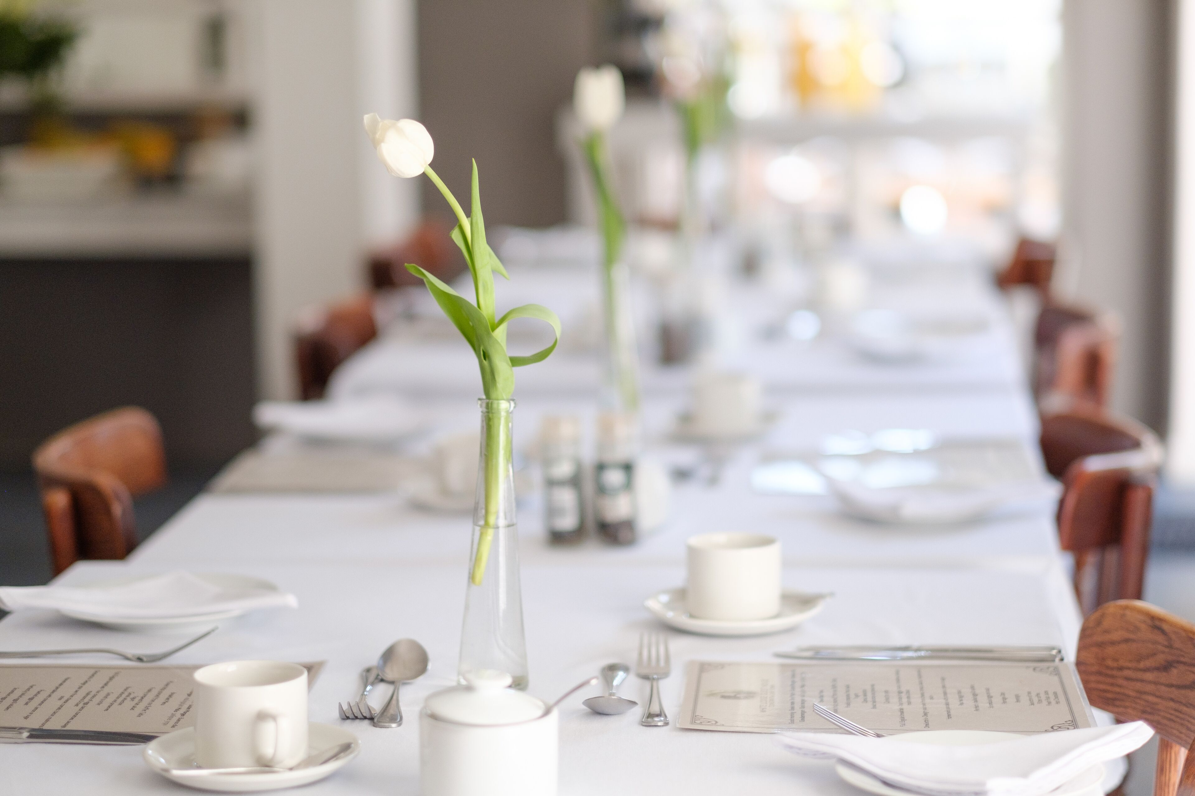 Una larga mesa de comedor dispuesta con vajilla blanca, un florero con una sola flor y un menú.