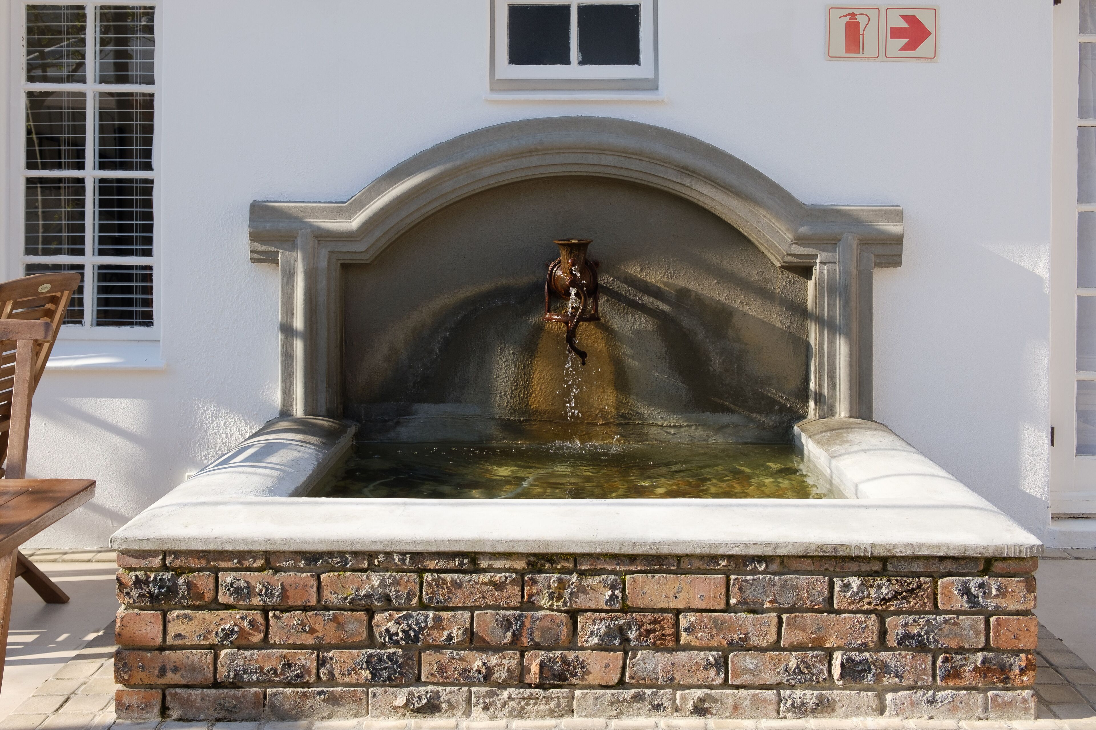 A small outdoor water feature with a fountain, surrounded by a brick base and a white wall.