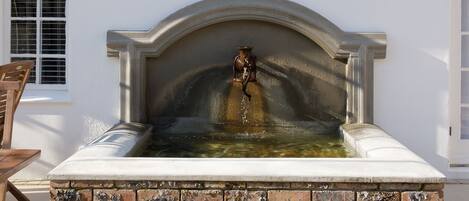 A small outdoor water feature with a fountain, surrounded by a brick base and a white wall.