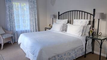 Una cama bien hecha con sábanas blancas y motivos florales azules, flanqueada por dos mesitas de noche con lámparas, una silla de mimbre y un gran ventanal con cortinas traslúcidas.