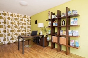 A room with a wooden desk, a laptop, a lamp, a bookshelf with magazines, and a patterned curtain. - Aarangi Tui Motel (Paihia)