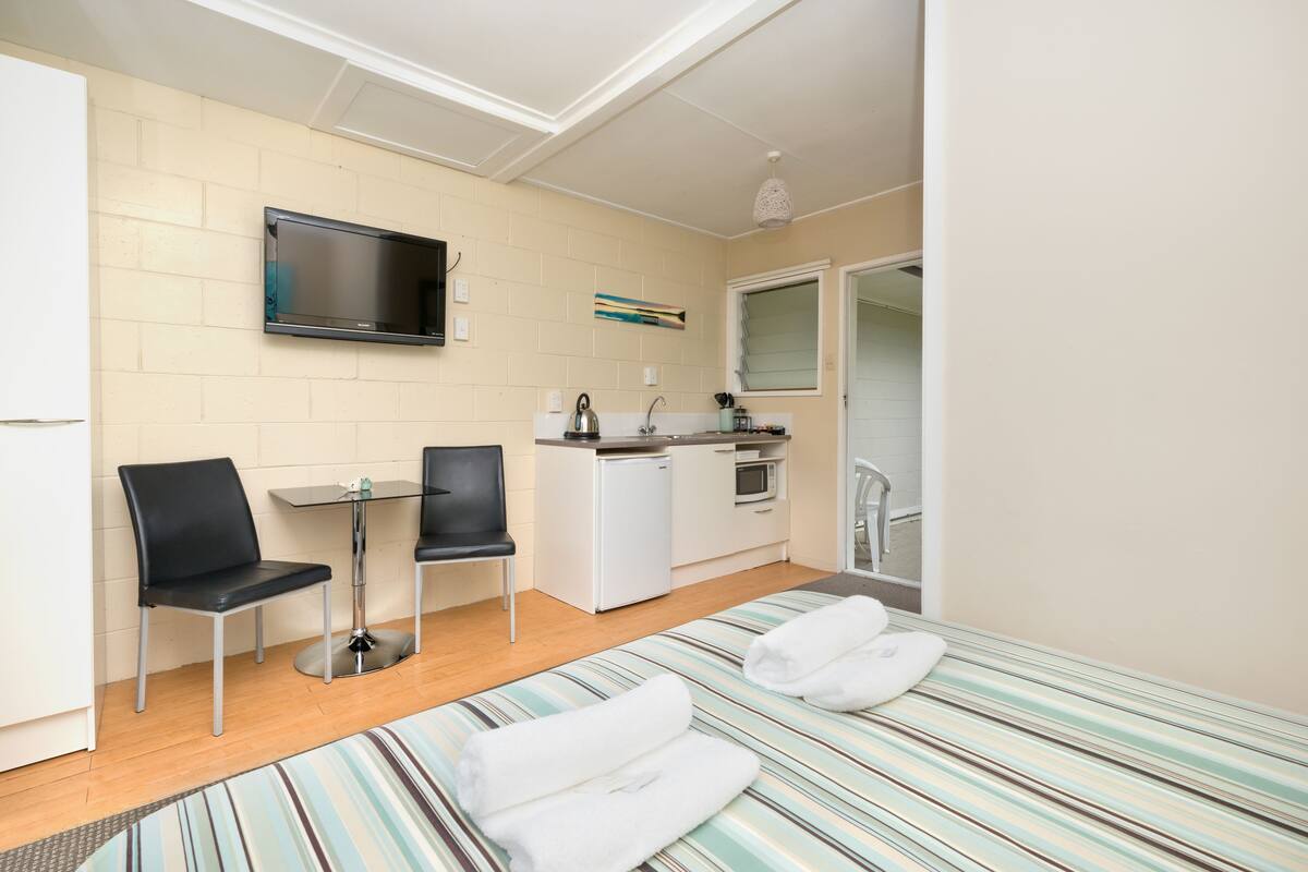 Una habitación compacta con cama, una kitchenette pequeña, un televisor montado en la pared y una ventana.