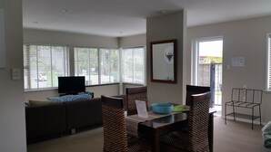 A living room with a sofa, a TV, a dining table with chairs, and large windows with blinds. - Aarangi Tui Motel (Paihia)