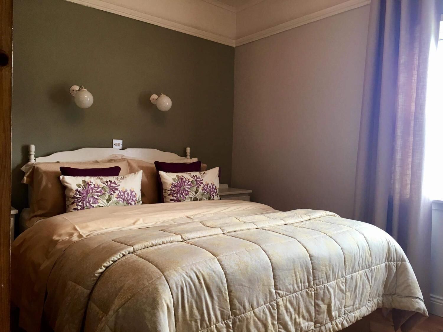Una cama bien hecha con colcha color beige, dos apliques decorativos y una ventana con cortinas traslúcidas.