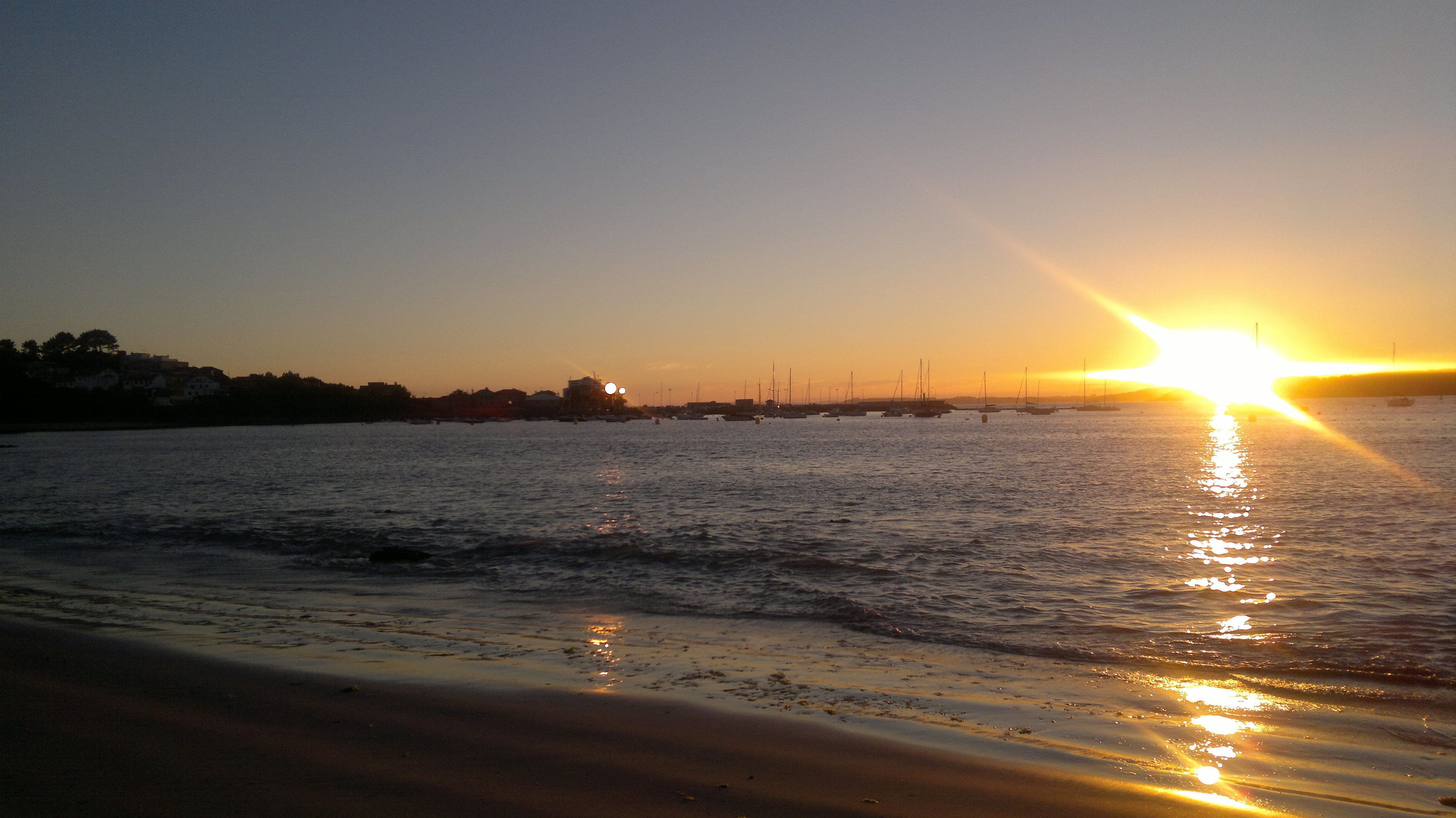 A beach at sunset with boats in the water and buildings in the background.