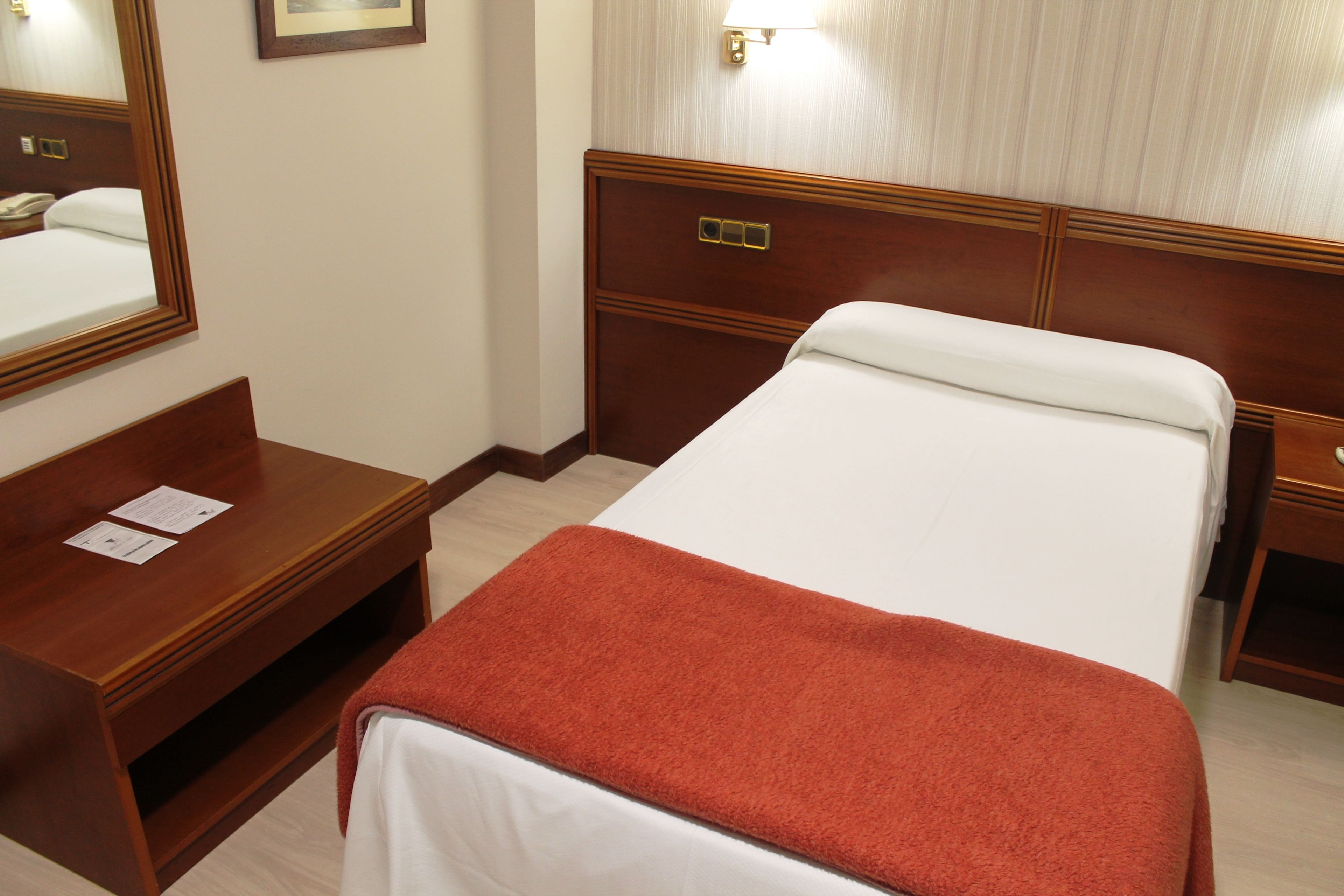 Una habitación de hotel con una cama, una mesita de noche de madera, un escritorio y un espejo.