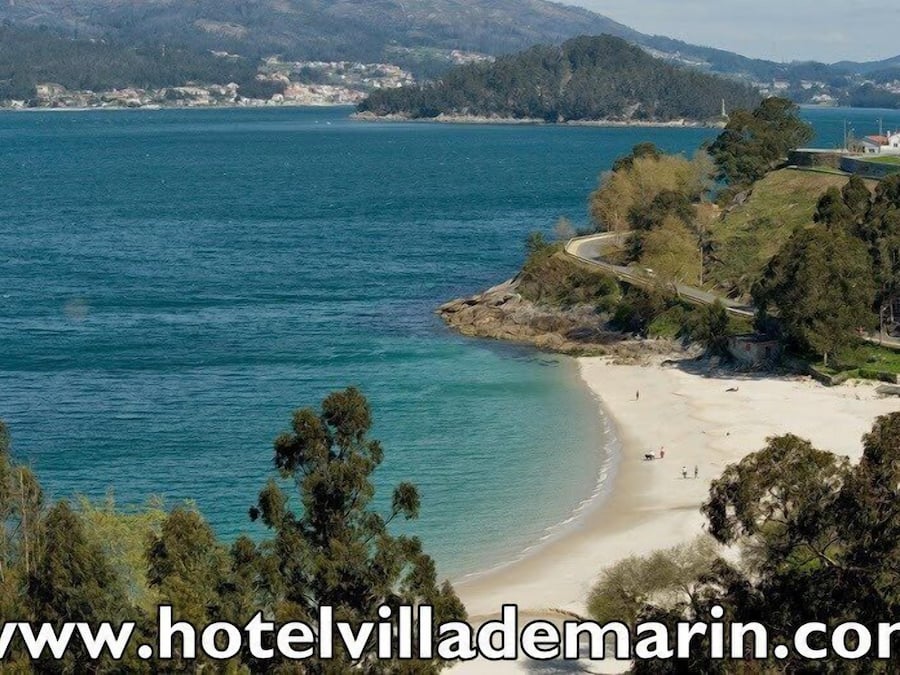 Vista costera con playa, una carretera sinuosa y una colina con edificios.