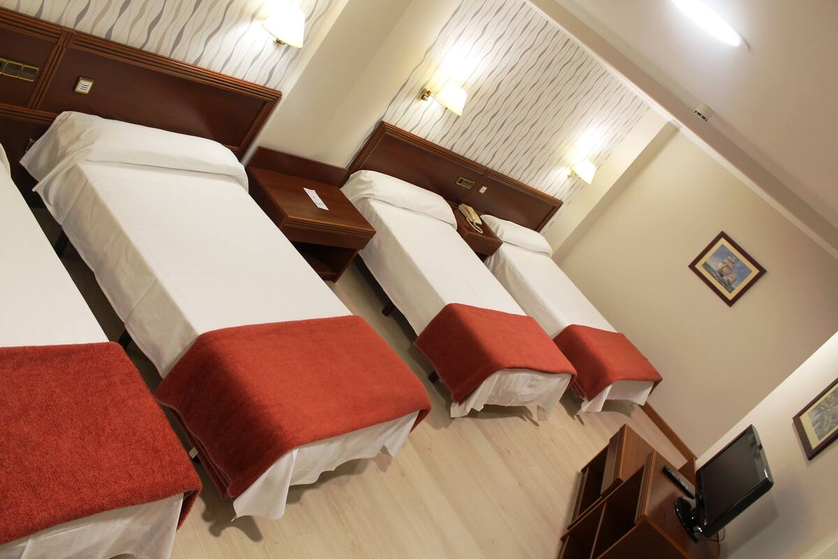 Une chambre d’hôtel avec trois lits simples, chacun recouvert d’une couverture rouge, dotée d’une tête de lit en bois et de tables de chevet avec des lampes.