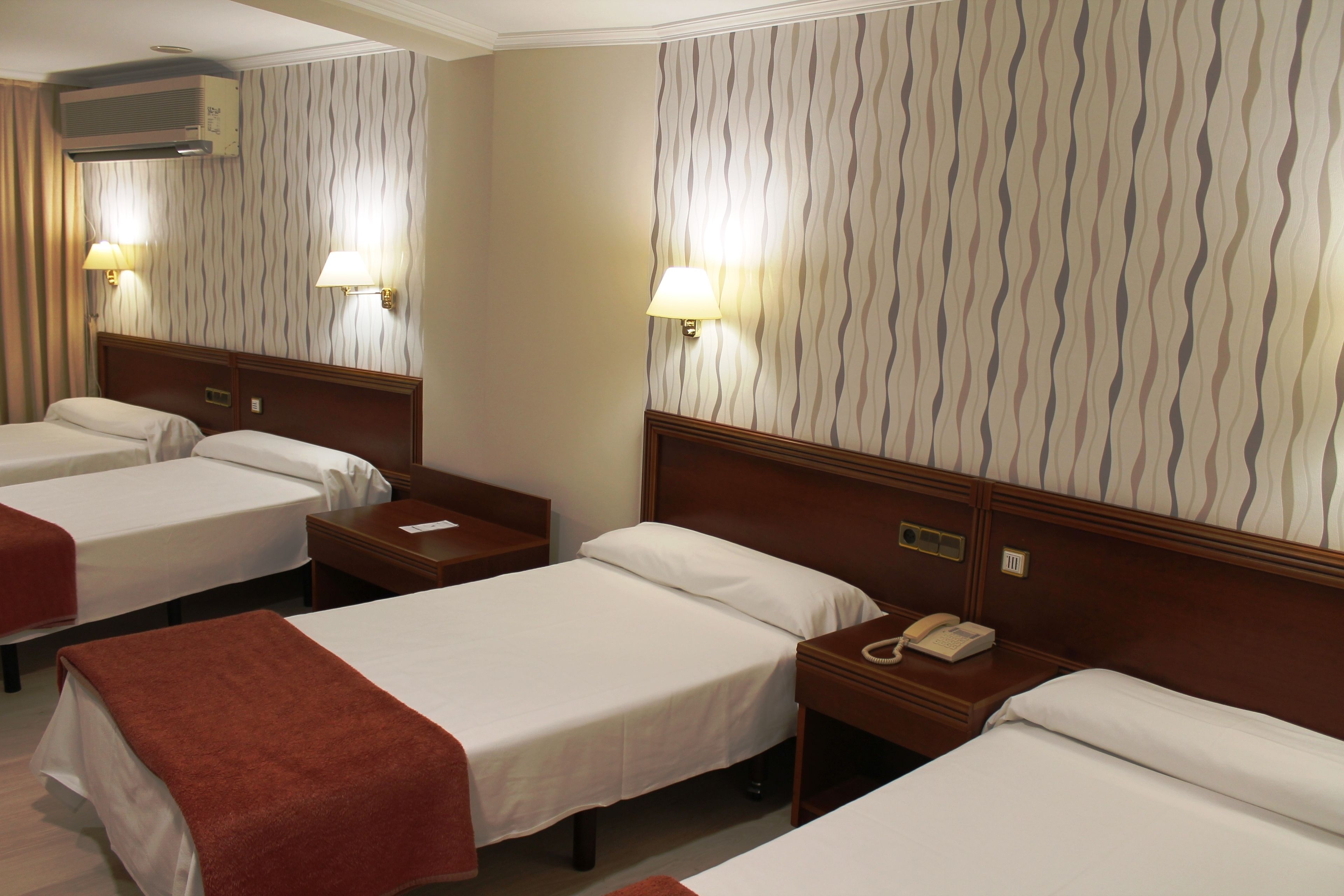Une chambre d’hôtel avec trois lits simples, chacun doté d’une tête de lit en bois et de tables de chevet. Des lampes murales sont fixées de chaque côté des lits, et un appareil de climatisation est installé au-dessus du mur gauche.
