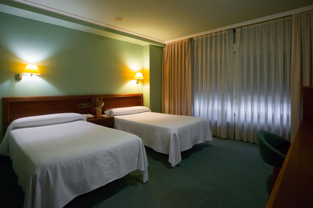 Une chambre d’hôtel avec deux lits simples, une tête de lit en bois, une table de chevet avec une lampe et une grande fenêtre avec des rideaux.