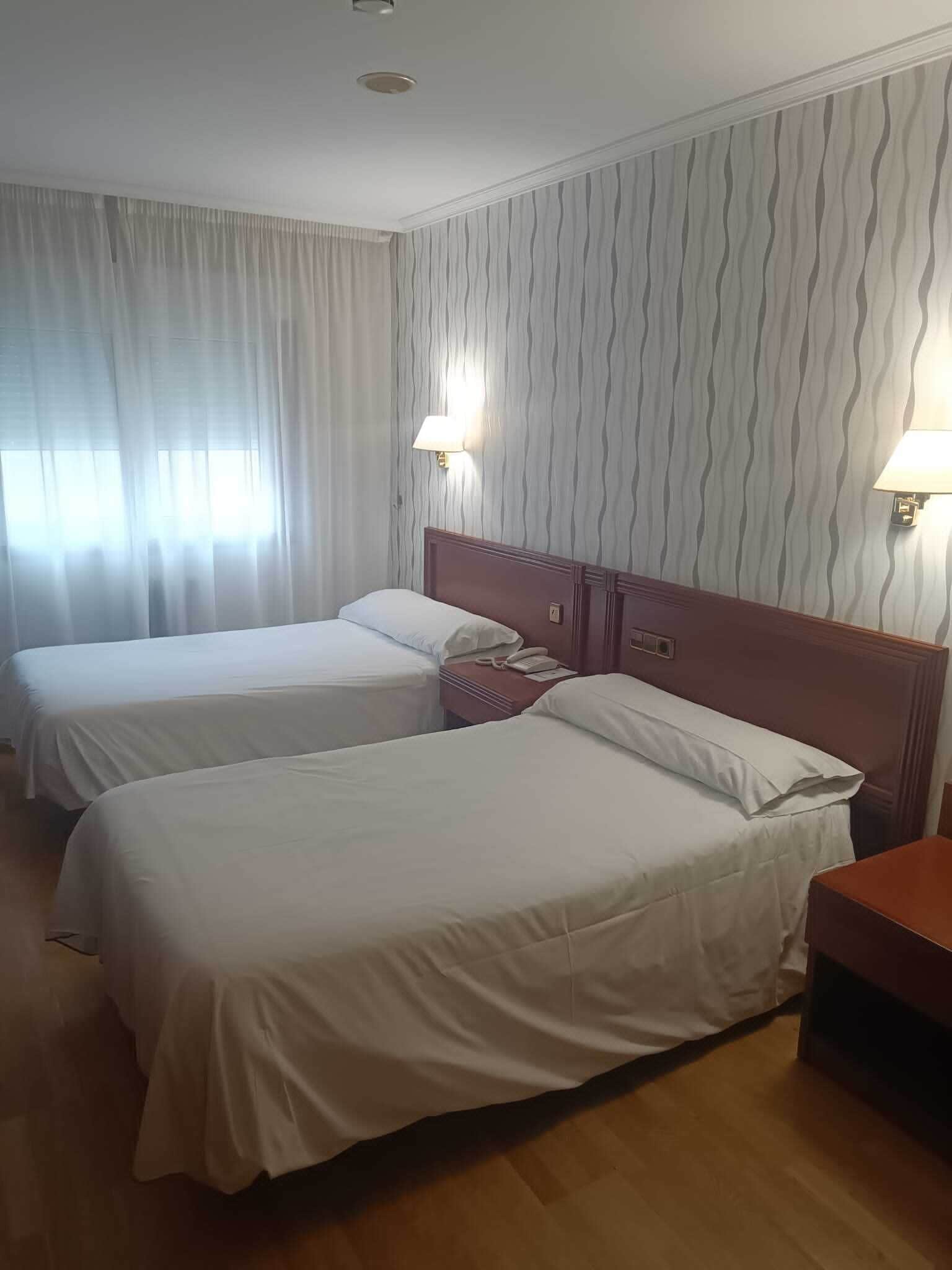 A hotel room with two beds, a wooden headboard, a nightstand, and a lamp.