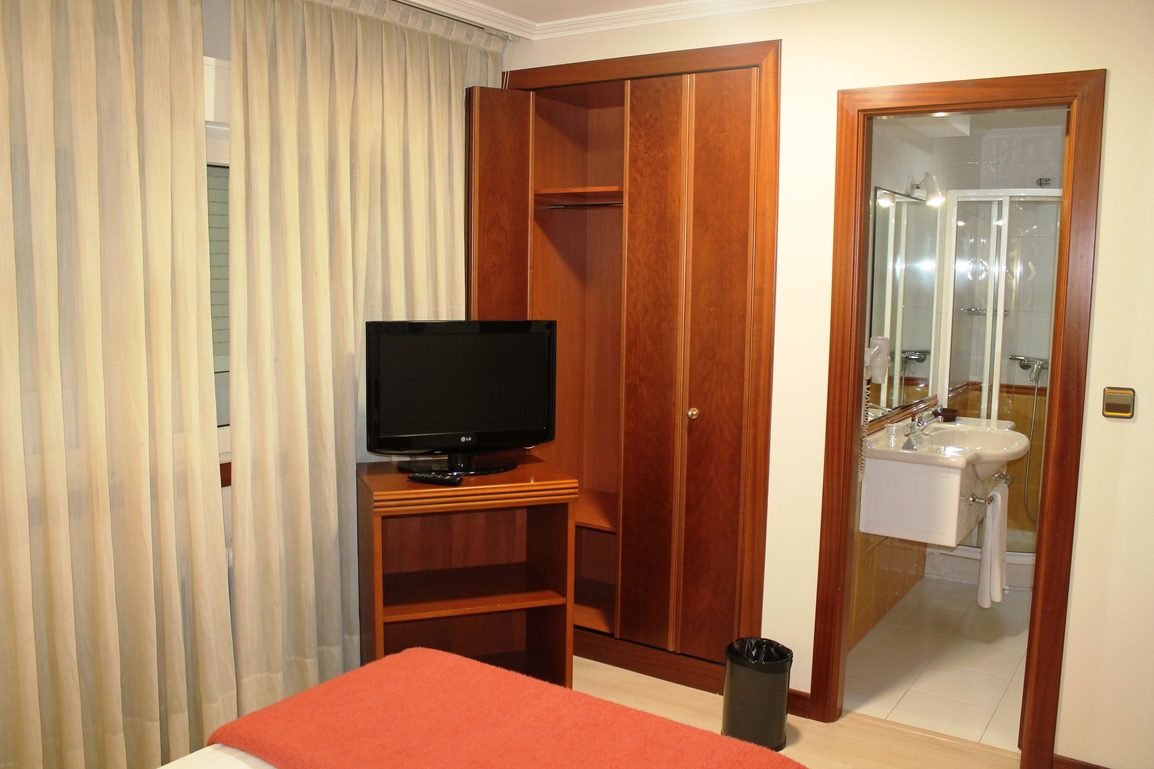 A hotel room with a bed, a television, a wardrobe, a bathroom with a sink and mirror, and a trash bin.