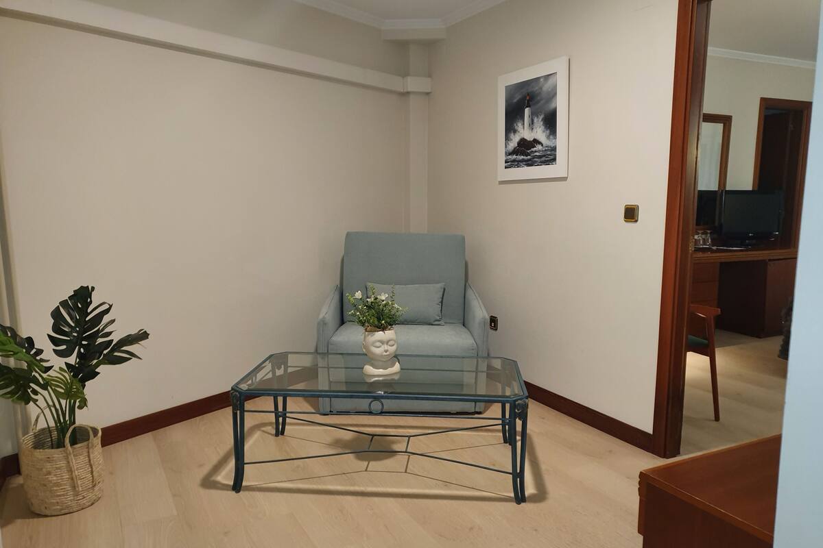 Un salon avec un fauteuil bleu clair, une table basse en verre surmontée d’un vase blanc et un tableau encadré accroché au mur.