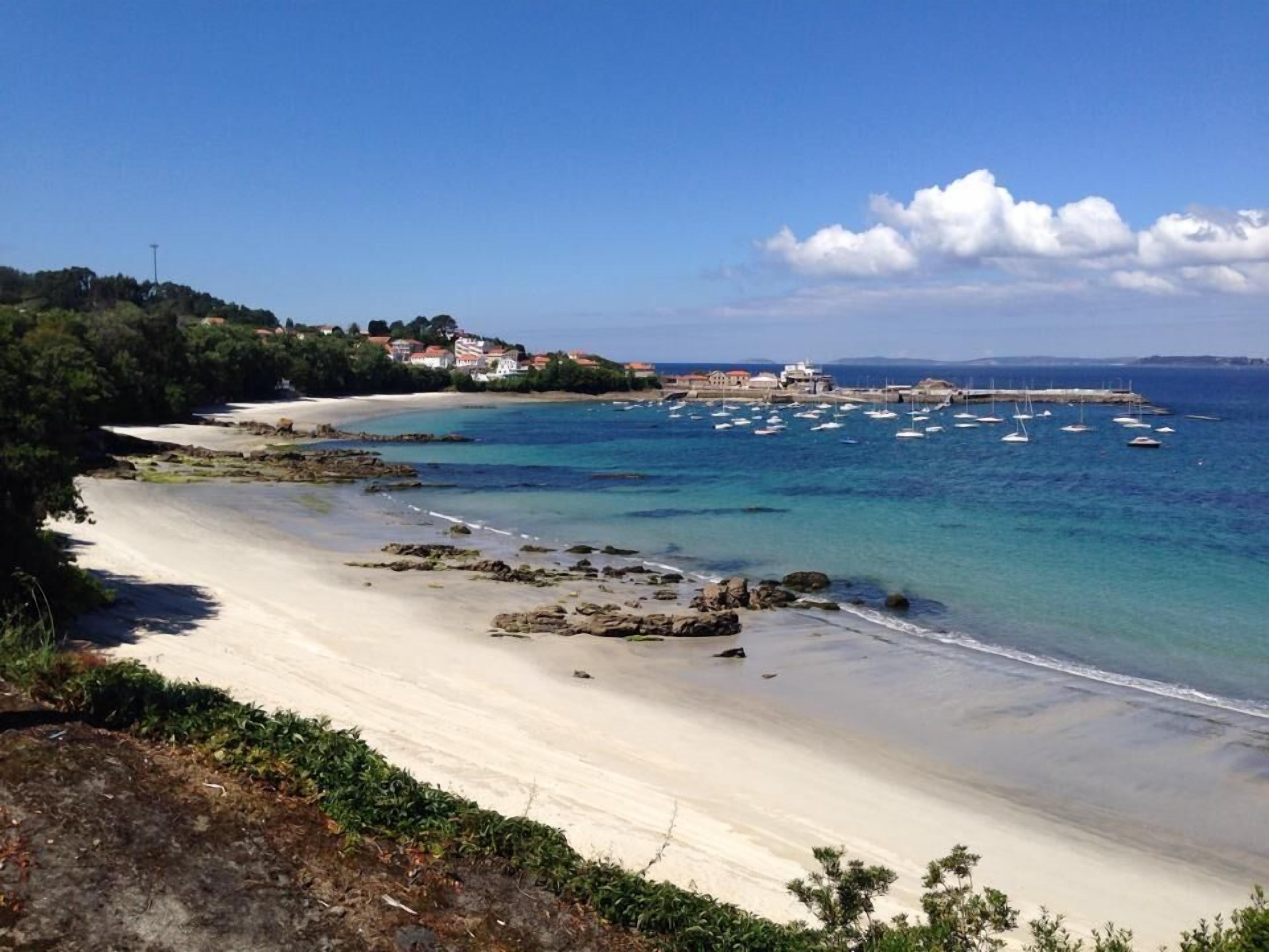 Uma praia costeira com águas cristalinas, areal arenoso e uma pequena cidade ao fundo.