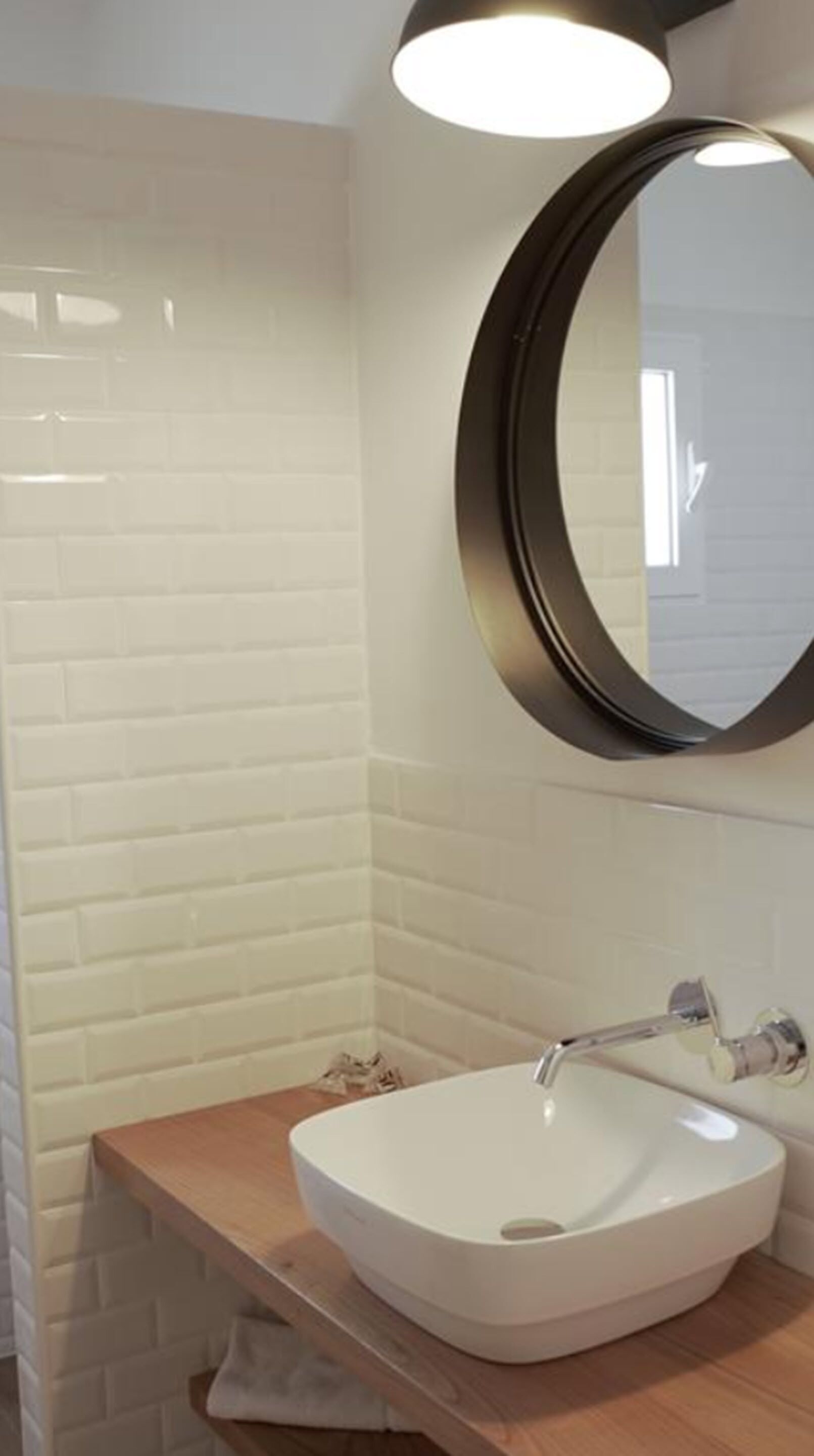 Une salle de bain avec des carreaux de céramique blancs rectangulaires, un meuble-lavabo en bois, un lavabo blanc et un miroir rond avec un cadre noir.