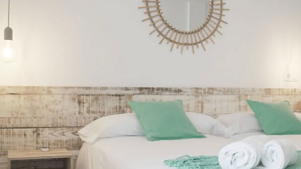 Een net opgemaakt bed met witte beddengoed en groene kussens, een ronde wanddecoratie en een klein nachtkastje met een lamp.